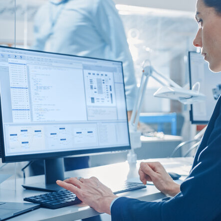 Back View of Team of Technology Engineers Working on Desktop Computers in Bright Office. Screens Show IDE / CAD Software, Implementation of Machine Learning, Neural Networking and Cloud Computing