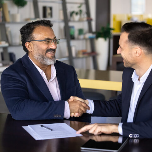 Happy business project buyer and seller men shaking hands, sitting at work table with documents, meeting for cooperation, partnership, getting agreement about investment. Vertical shot